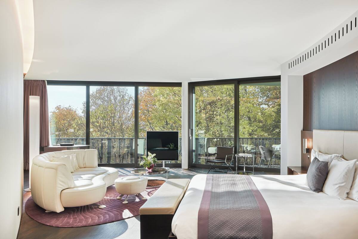 A modern hotel room with a large bed, a sofa, a TV, and a balcony with outdoor seating.