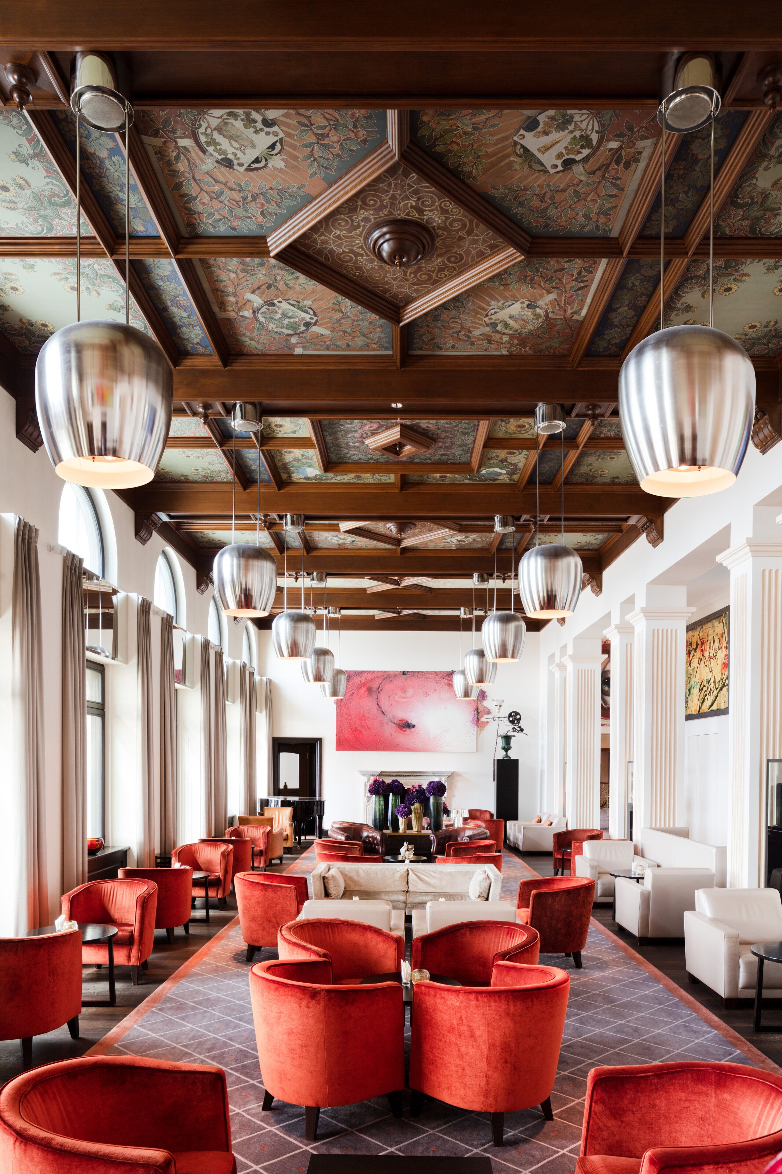A spacious hotel lobby with red upholstered chairs, a long table, and a large painting on the wall.