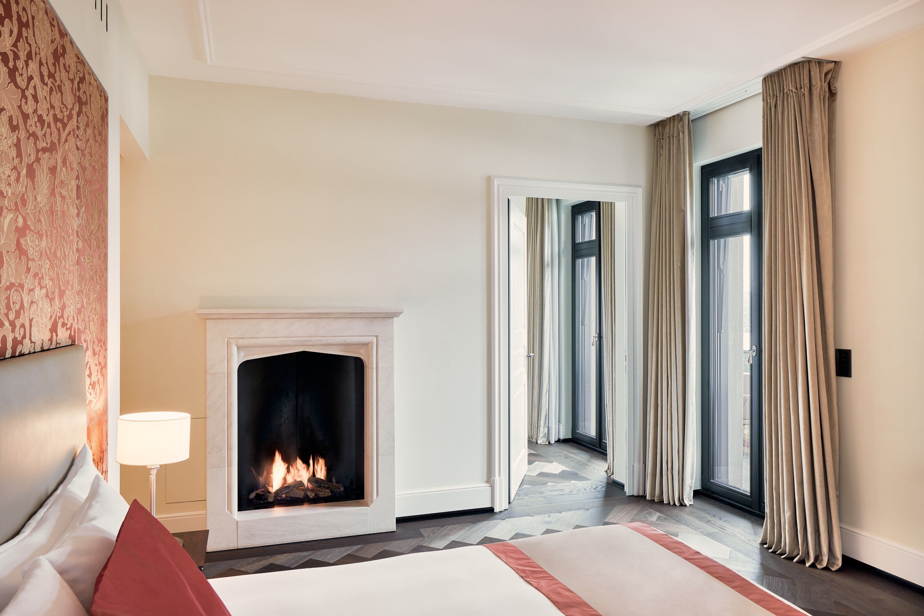 A hotel room with a fireplace, a bed, a nightstand, and a view through a large window.