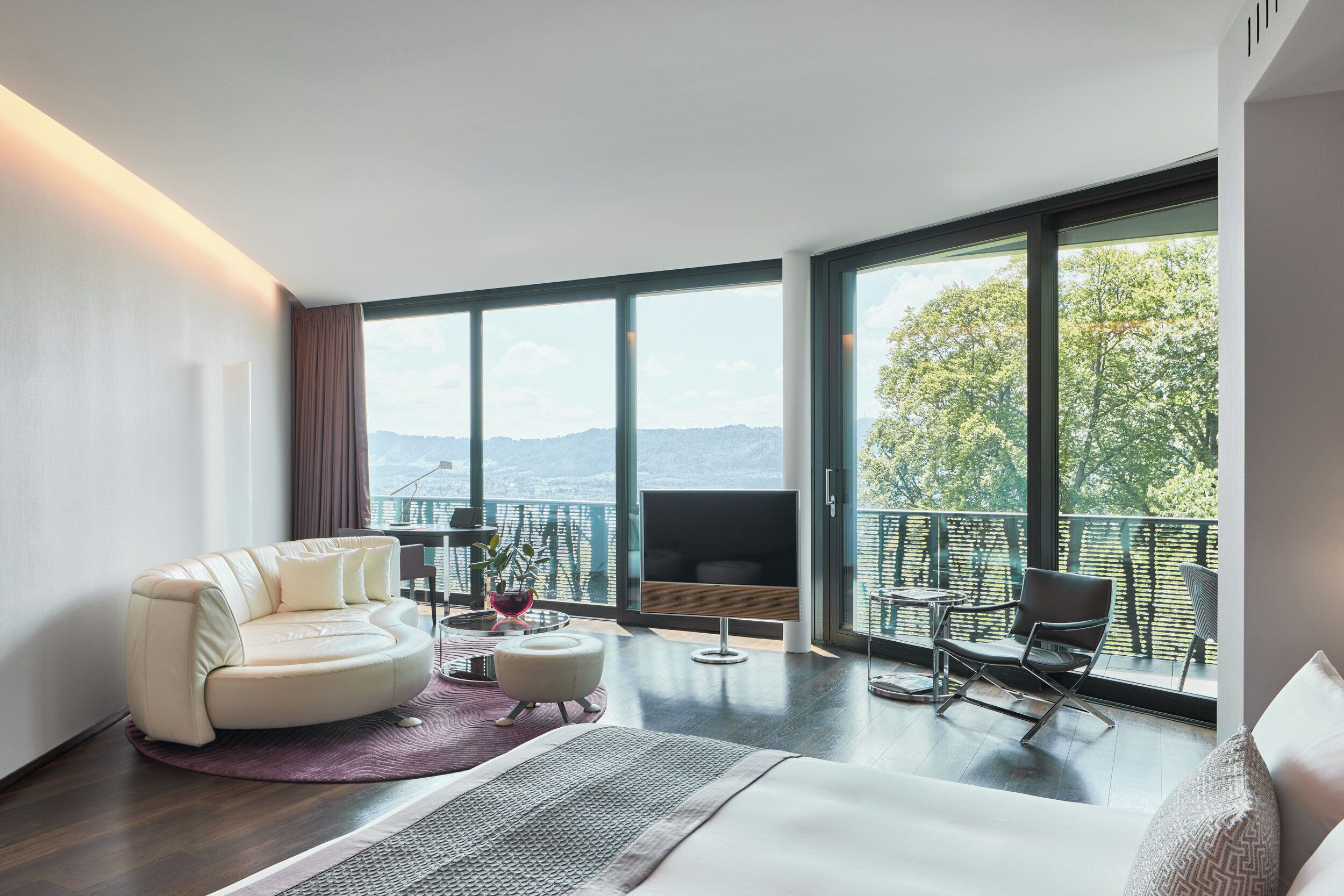 A modern hotel room with a large bed, a sofa, a TV, and a balcony with a view of the mountains.