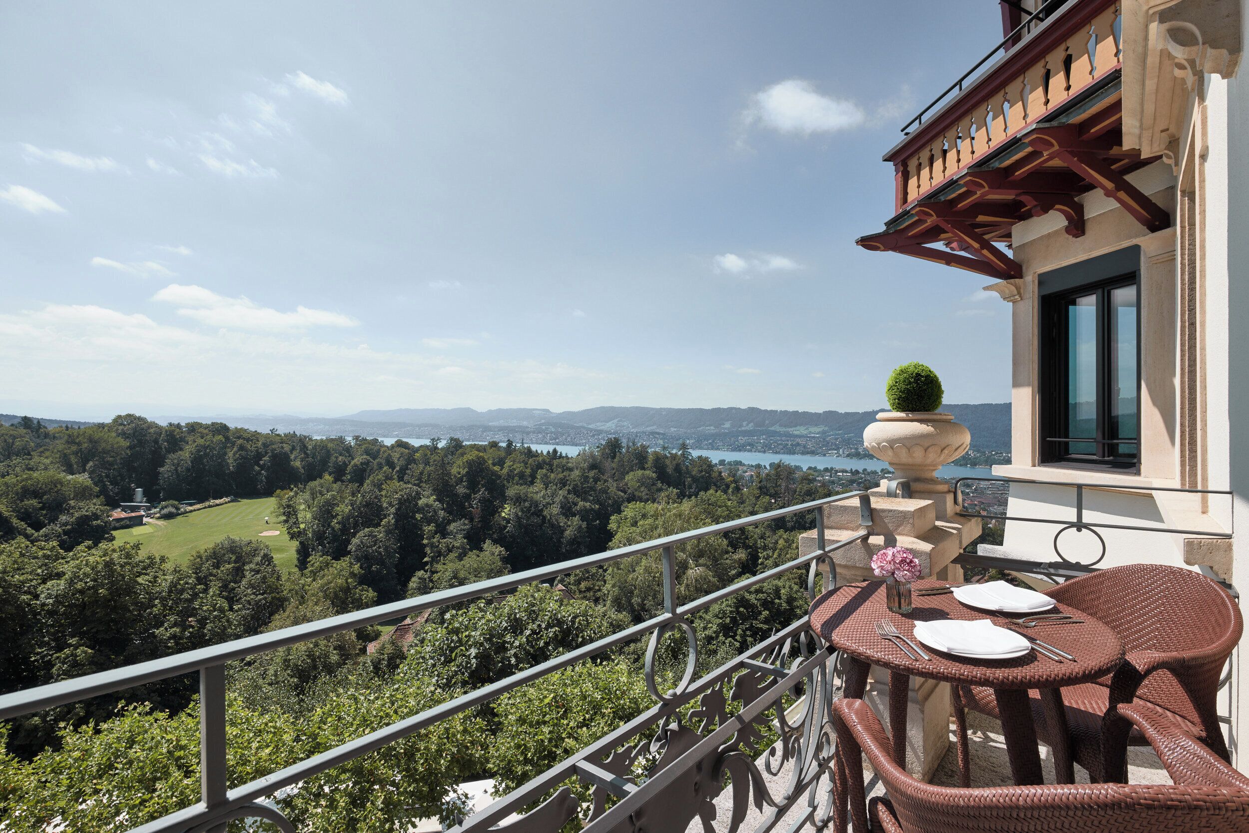 A balcony with wicker furniture, a vase with flowers, and a view of a lake and forest.