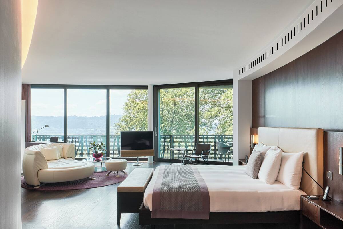 A modern hotel room with a large bed, a sofa, a TV, and a balcony with a view of trees and mountains.