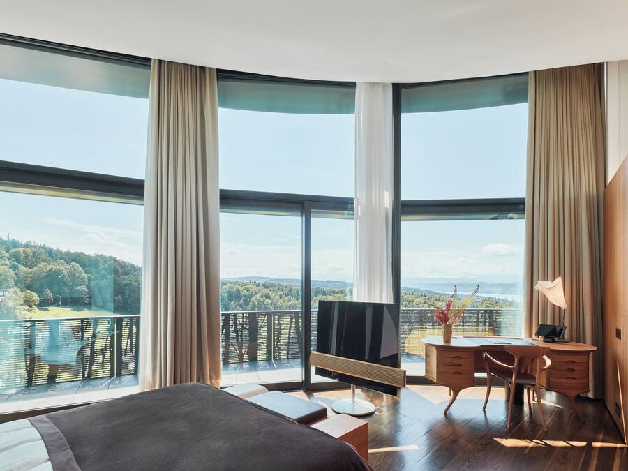 A modern bedroom with a large bed, a desk, and a TV. It features large windows offering a view of a forested area.