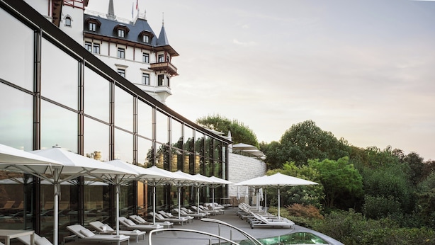 Un área de piscina moderna al aire libre con sillones de descanso y sombrillas, rodeada de exuberante vegetación y un edificio grande con una torre distintiva.