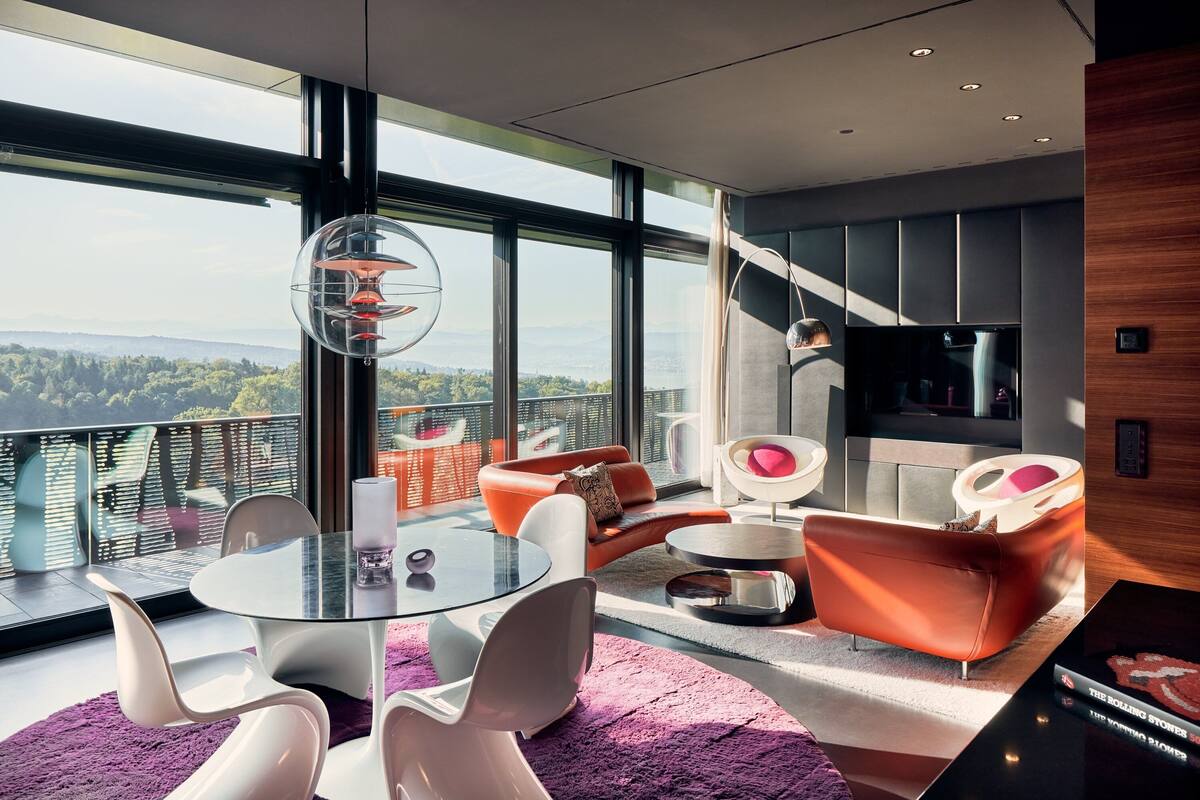 A modern living room with a glass table, white chairs, and orange sofas. A large window offers a view of the city and mountains.