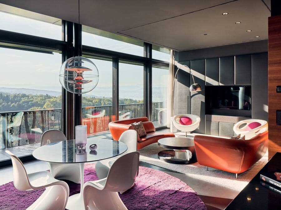 Una sala de estar moderna con una mesa de vidrio, sillas blancas y sofás naranjas. Un ventanal ofrece vistas a la ciudad y a las montañas.