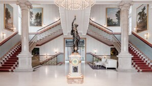 Un vestíbulo imponente con una gran araña, columnas de mármol, una estatua y una escalera con peldaños cubiertos de alfombra roja.