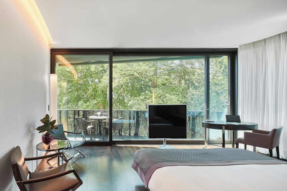 A modern hotel room with a large bed, a desk with a laptop, a TV, and a balcony with outdoor seating.