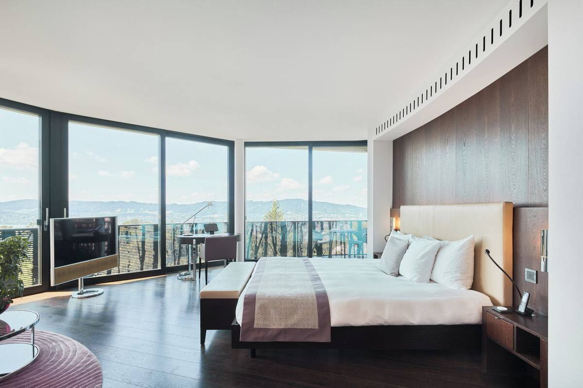A modern hotel room with a large bed, a TV, a balcony with a view of the city, and a desk with a chair.