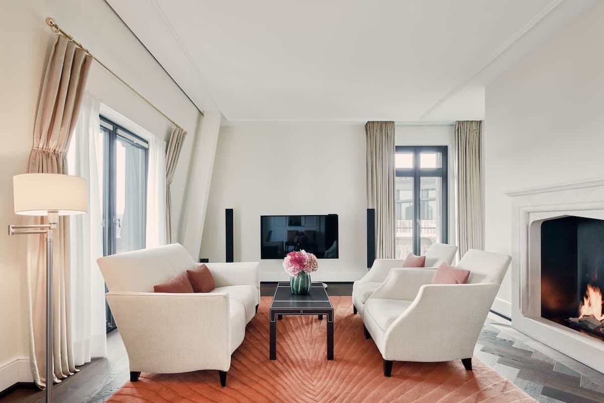 A modern living room with two white armchairs, a dark wooden coffee table, and a vase of flowers. A flat-screen TV is mounted on the wall, and a fireplace is visible on the right.
