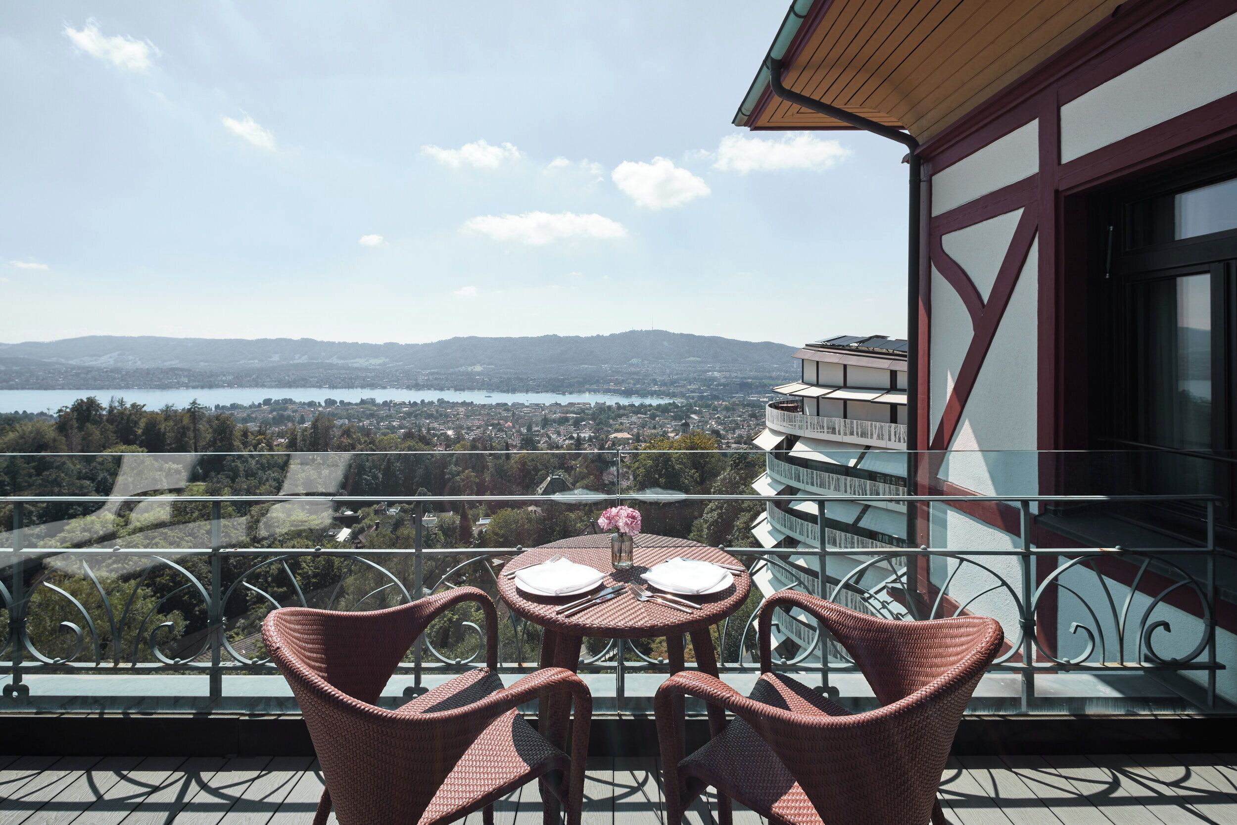 A balcony with wicker chairs and a table, overlooking a scenic view of a lake and distant mountains.