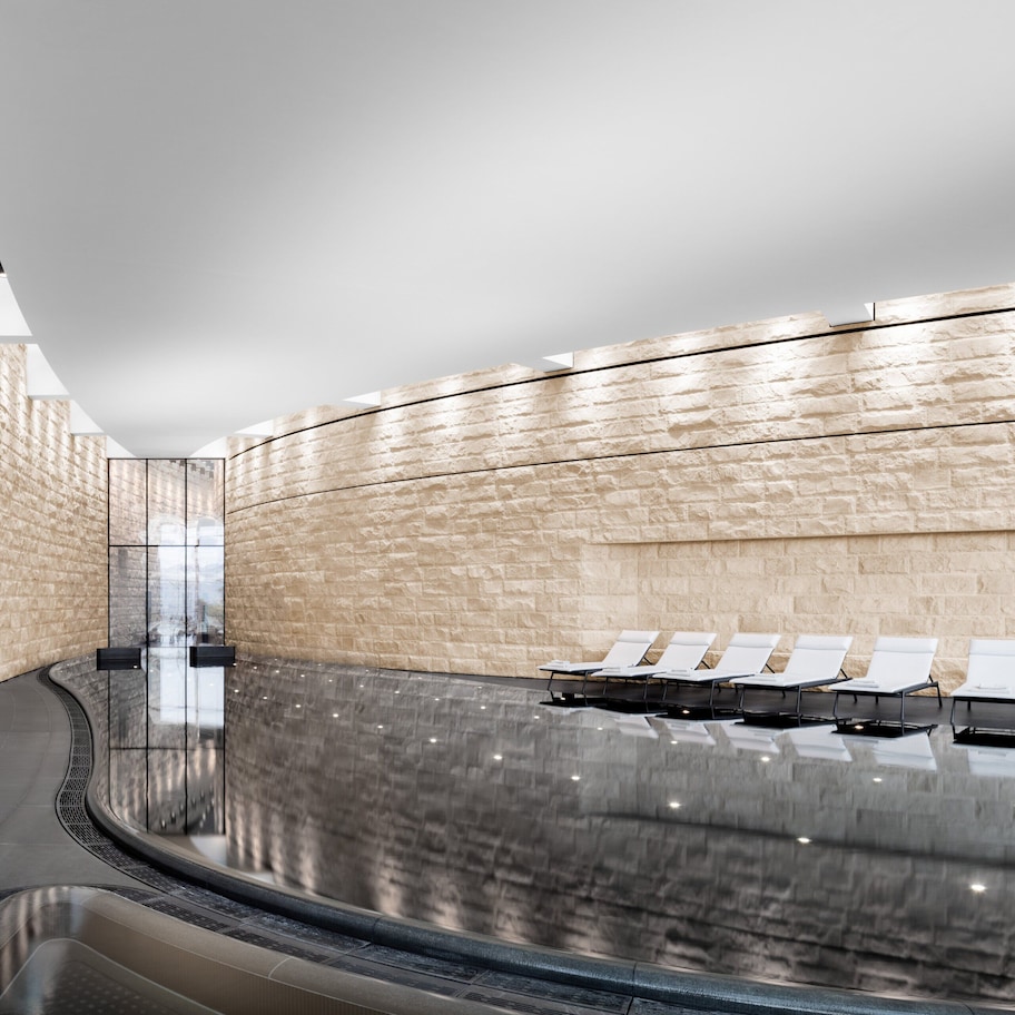 A modern indoor pool area with a curved glass wall, seating along the pool, and a stone wall on one side.
