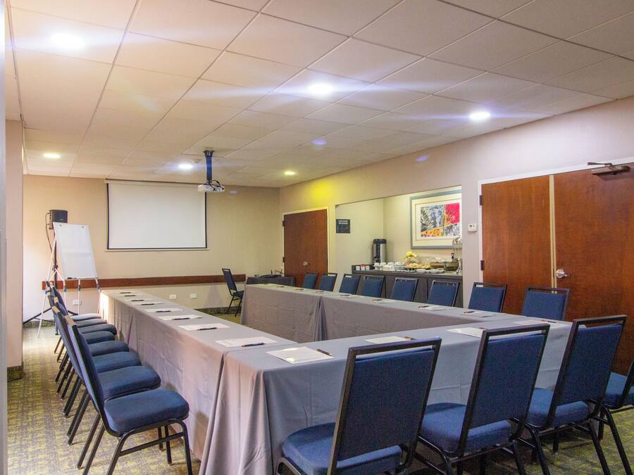 Un salón de conferencias con una larga mesa, sillas, un proyector y una pantalla.