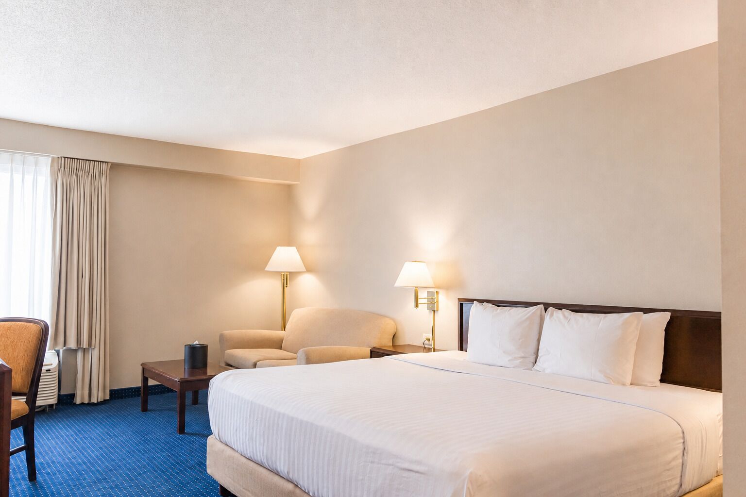Habitación de hotel con una cama grande, dos lámparas de noche, una silla, una mesita y un ventanal con cortinas.