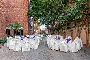 Montaje para una boda al aire libre con mesas y sillas dispuestas en filas, rodeado de edificios de ladrillo y follaje.