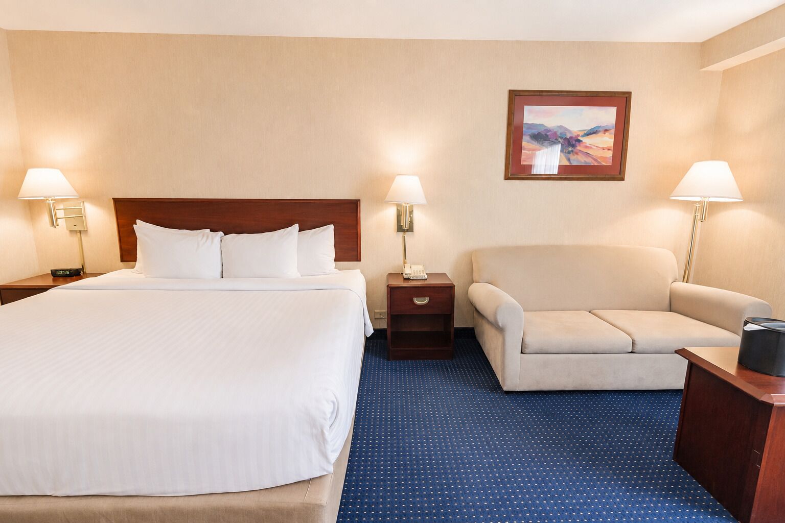 A hotel room with a large bed, two bedside tables with lamps, a sofa, and a painting on the wall.