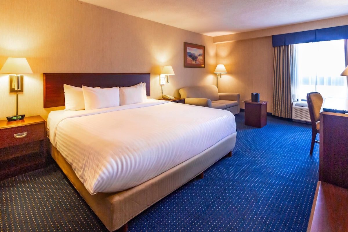 Habitación de hotel con una cama grande, dos mesitas de noche, un escritorio, una silla y un ventanal con cortinas.