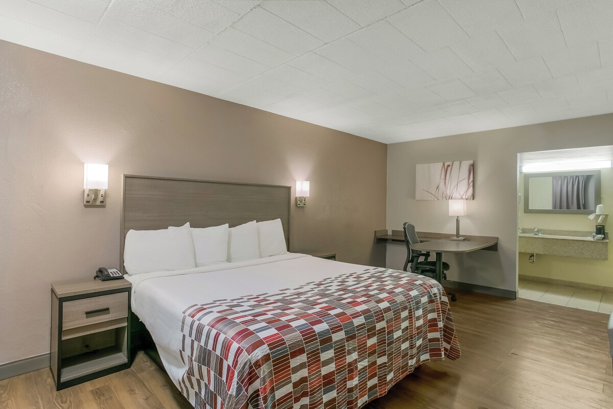 A hotel room with a bed, bedside table, lamp, desk, chair, and a bathroom with a mirror.