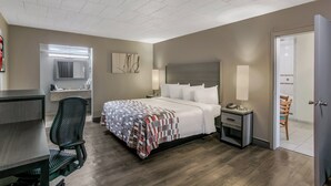 Quarto de hotel com cama, escrivaninha, cadeira, mesa de cabeceira, luminária e porta que dá para outro quarto.