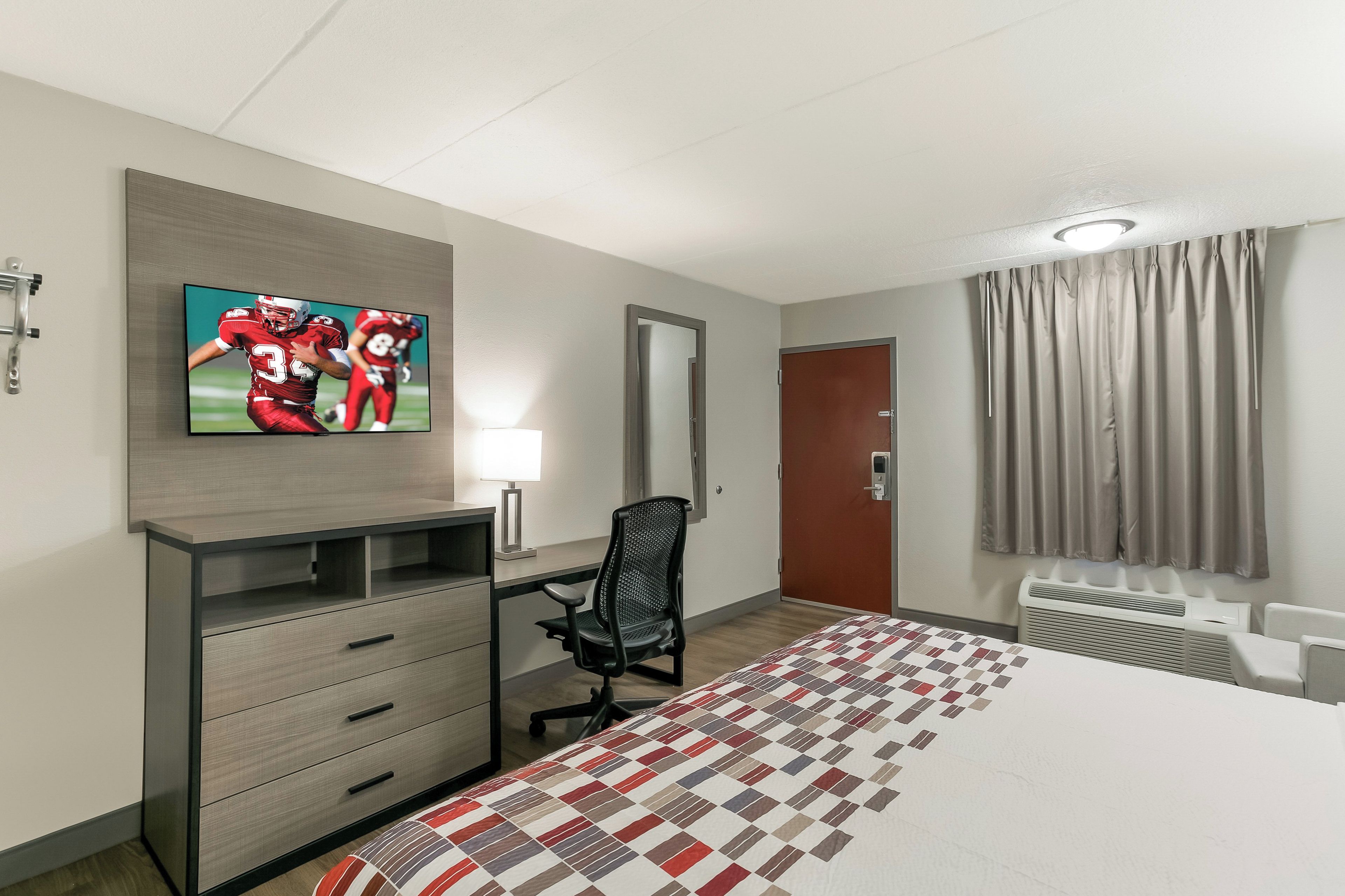 A hotel room with a bed, desk, chair, television, mirror, and a door.