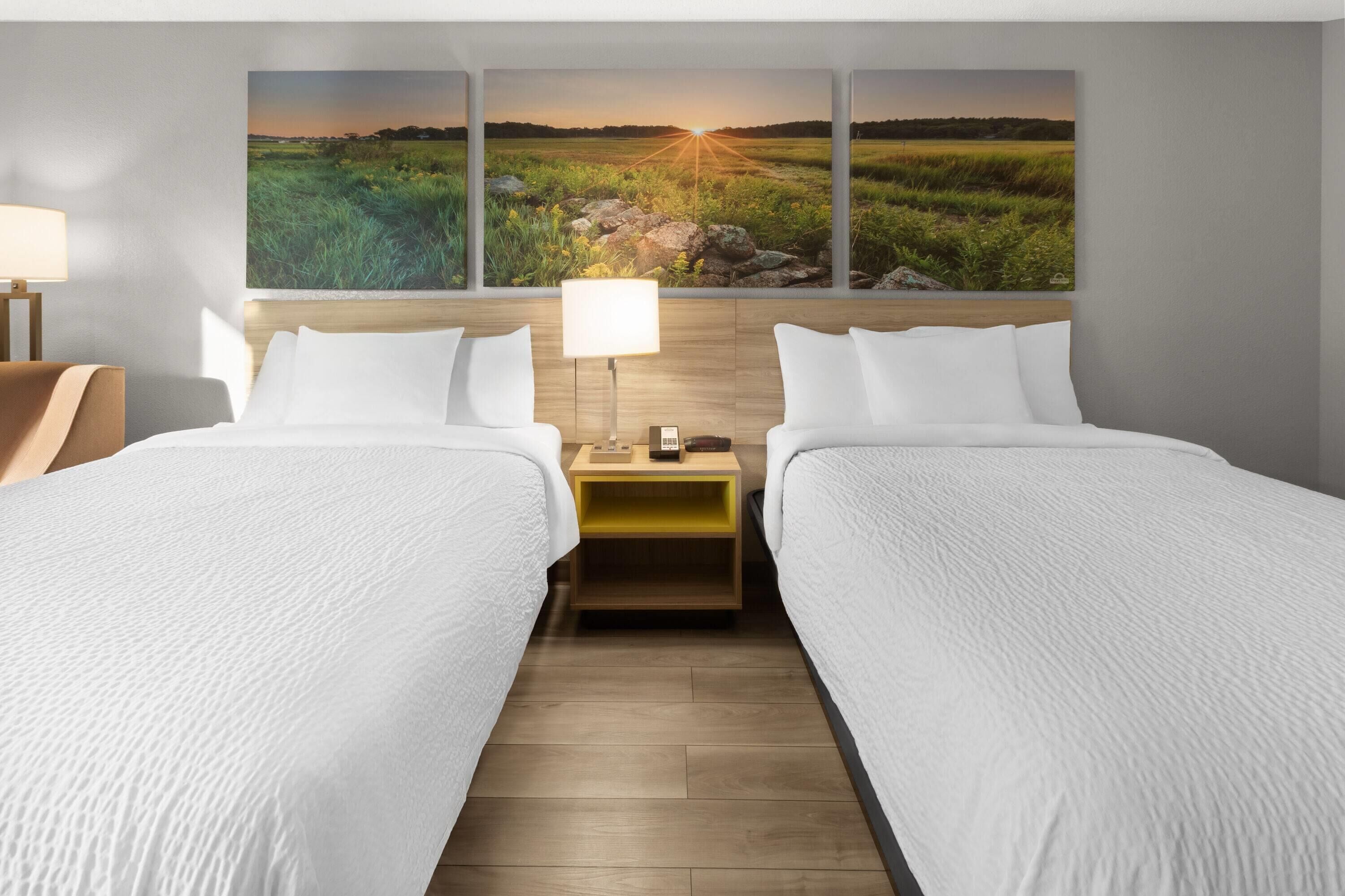 Habitación de hotel con dos camas, una mesita de noche de madera, una lámpara y un cuadro grande con un paisaje en la pared.