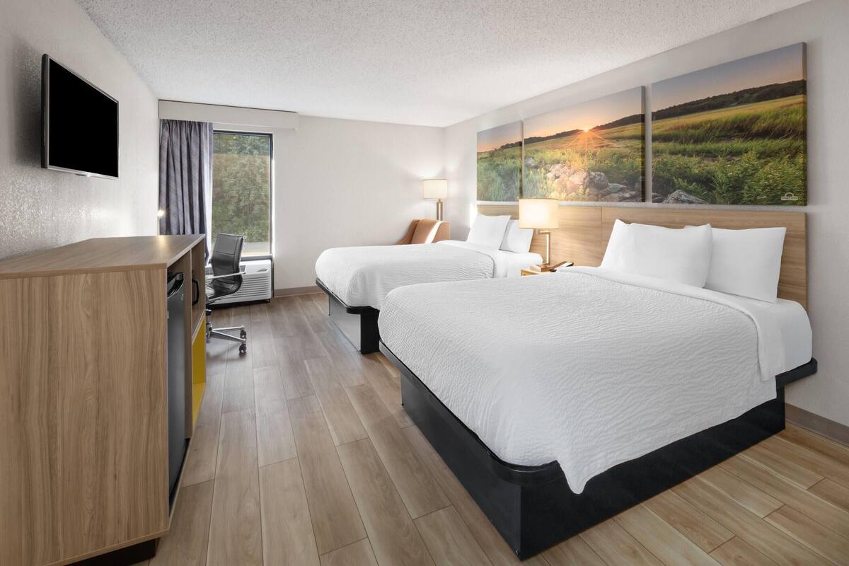 Hotelkamer met twee bedden, een bureau, een stoel, een televisie en een groot raam met gordijnen.