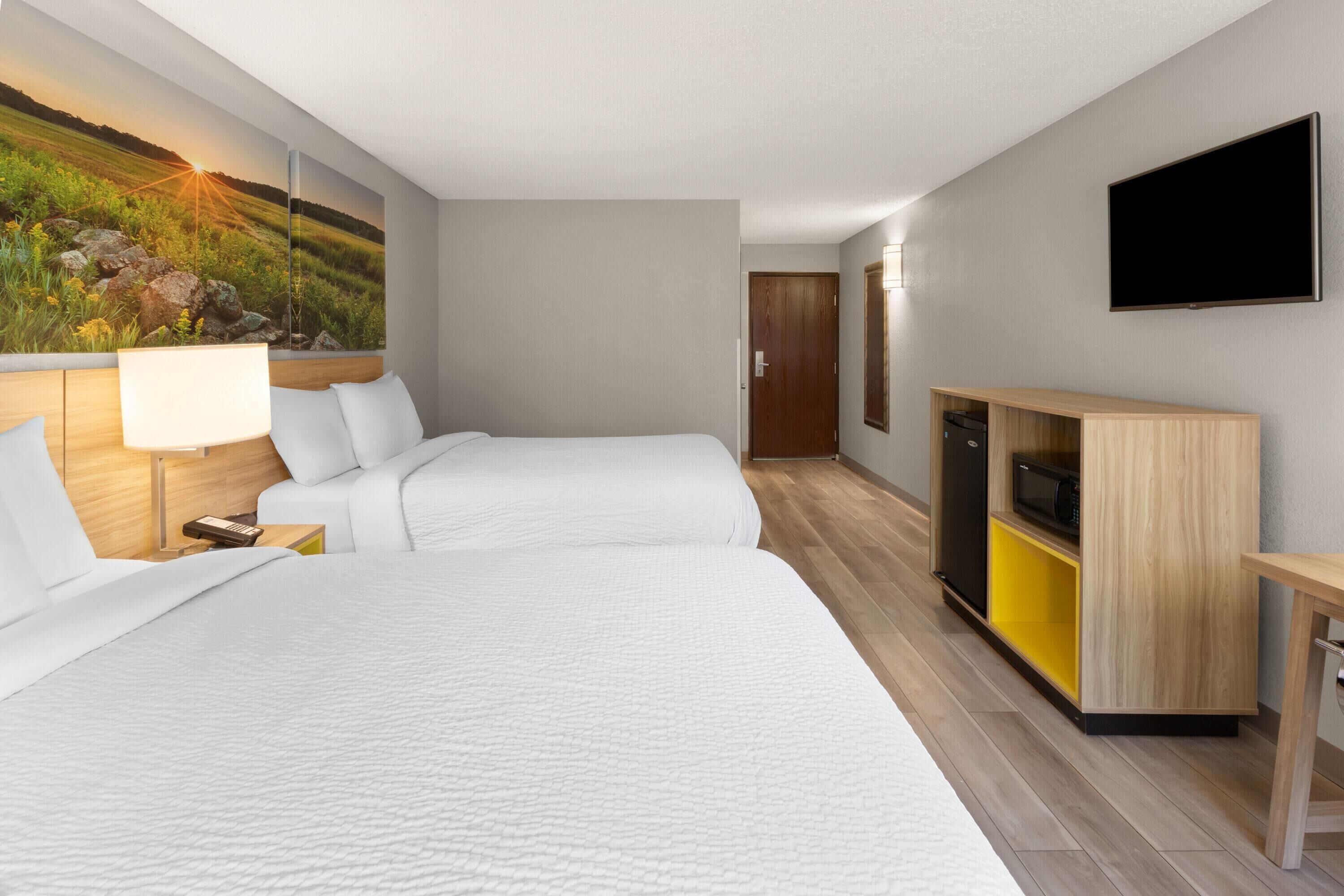 A hotel room with two beds, a TV, a wooden cabinet with a microwave, and a painting on the wall.