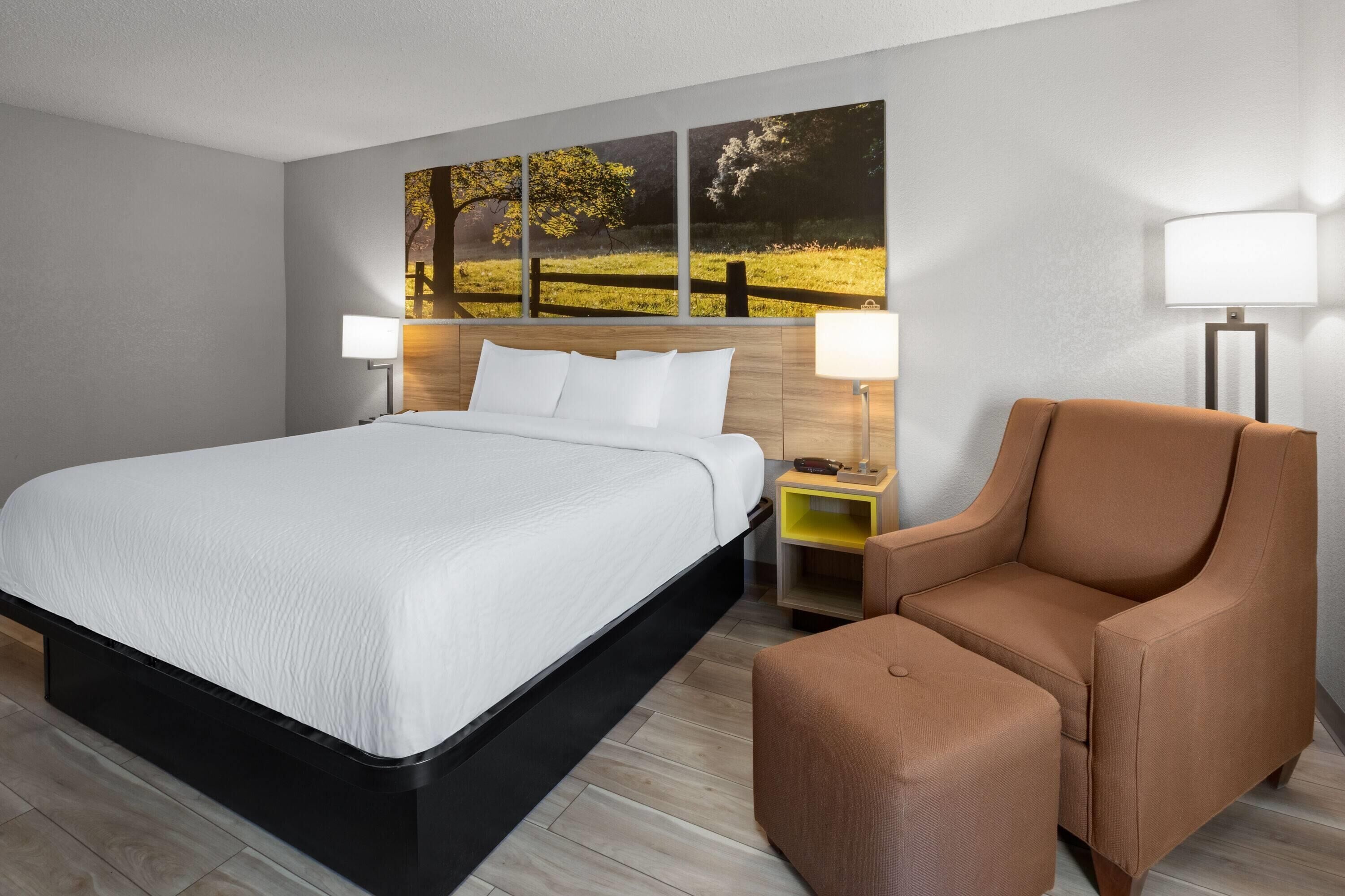 A modern hotel room with a large bed, a brown armchair, and a nightstand with a lamp.