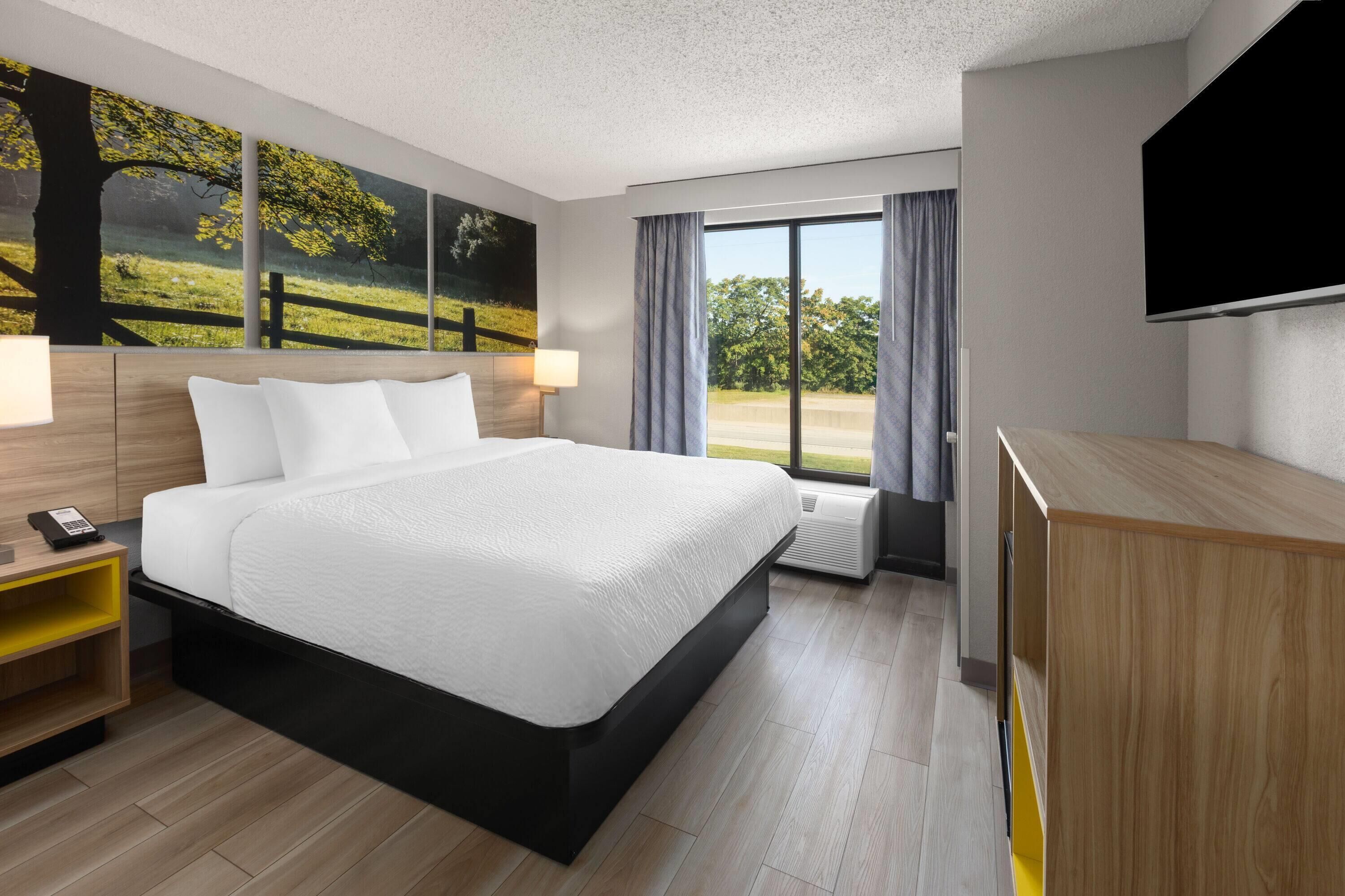 A modern hotel room with a large bed, a flat-screen TV, a desk, and a window with a view of greenery.