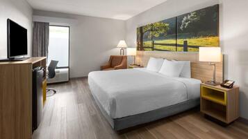 A hotel room with a bed, a desk with a computer, a chair, a lamp, and a painting on the wall.