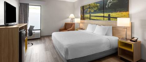 Quarto de hotel com uma cama, uma escrivaninha com computador, uma cadeira, um abajur e um quadro na parede.