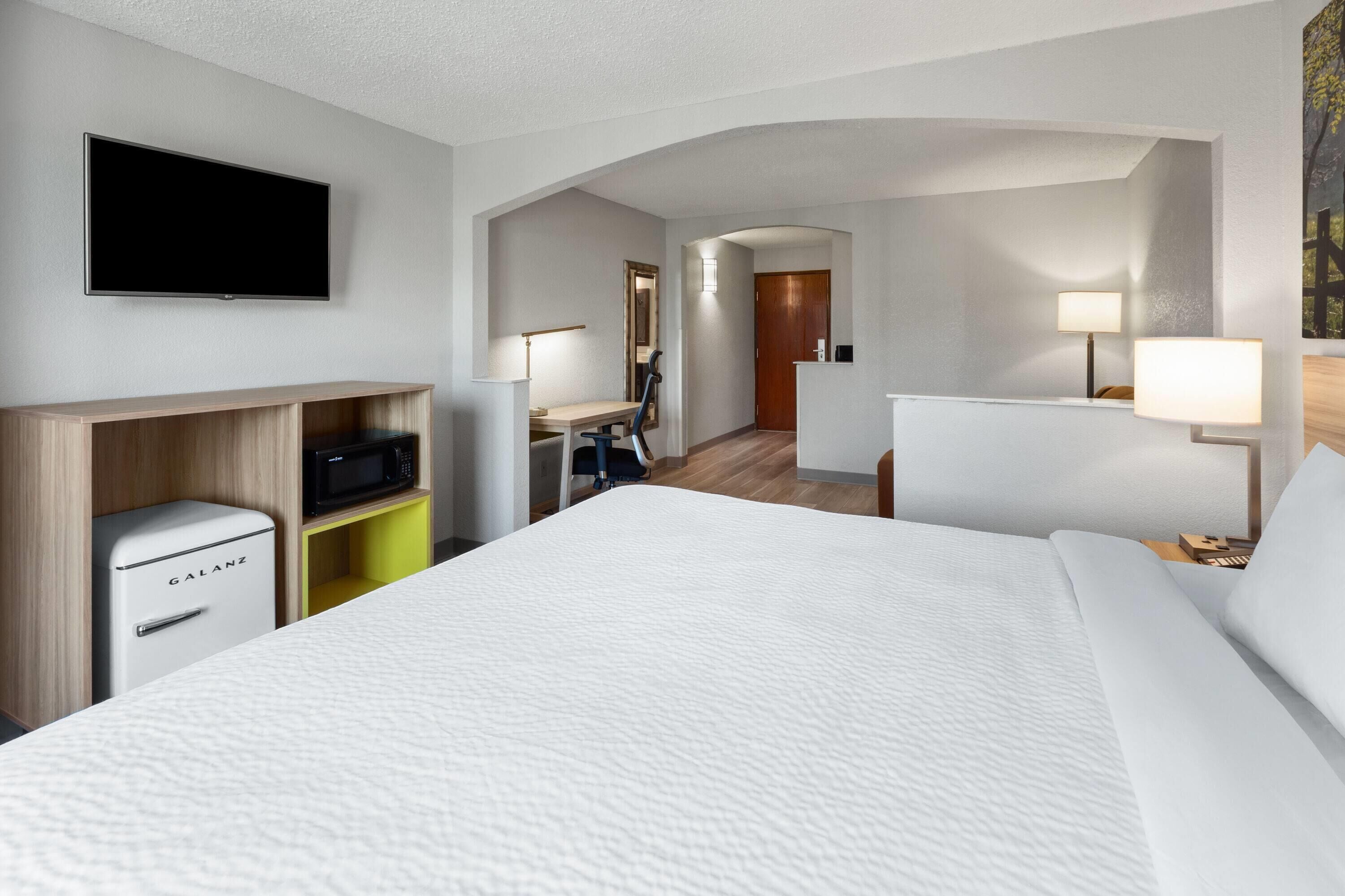 Habitación de hotel con cama, televisor de pantalla plana montado en la pared, escritorio con silla, microondas y refrigerador pequeño.