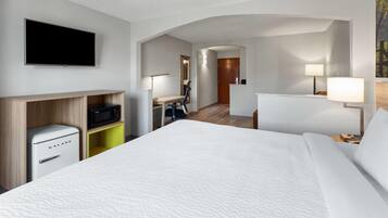 Quarto de hotel com cama, TV de tela plana na parede, escrivaninha com cadeira, micro-ondas e pequeno frigorífico.