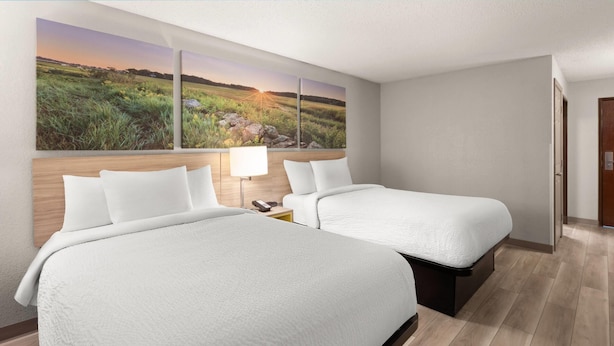 Ein Hotelzimmer mit zwei Betten, einem Holzkopfende, einem Nachttisch mit Lampe und einem großen Landschaftsbild an der Wand.