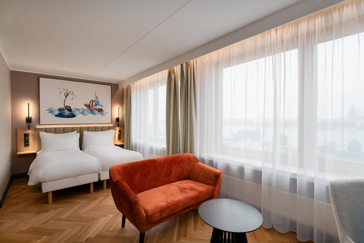 A hotel room with two beds, a red sofa, a small round table, and a large window with sheer curtains.