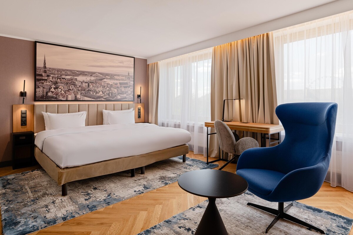 A hotel room with a bed, a desk, a chair, and a large window with curtains.