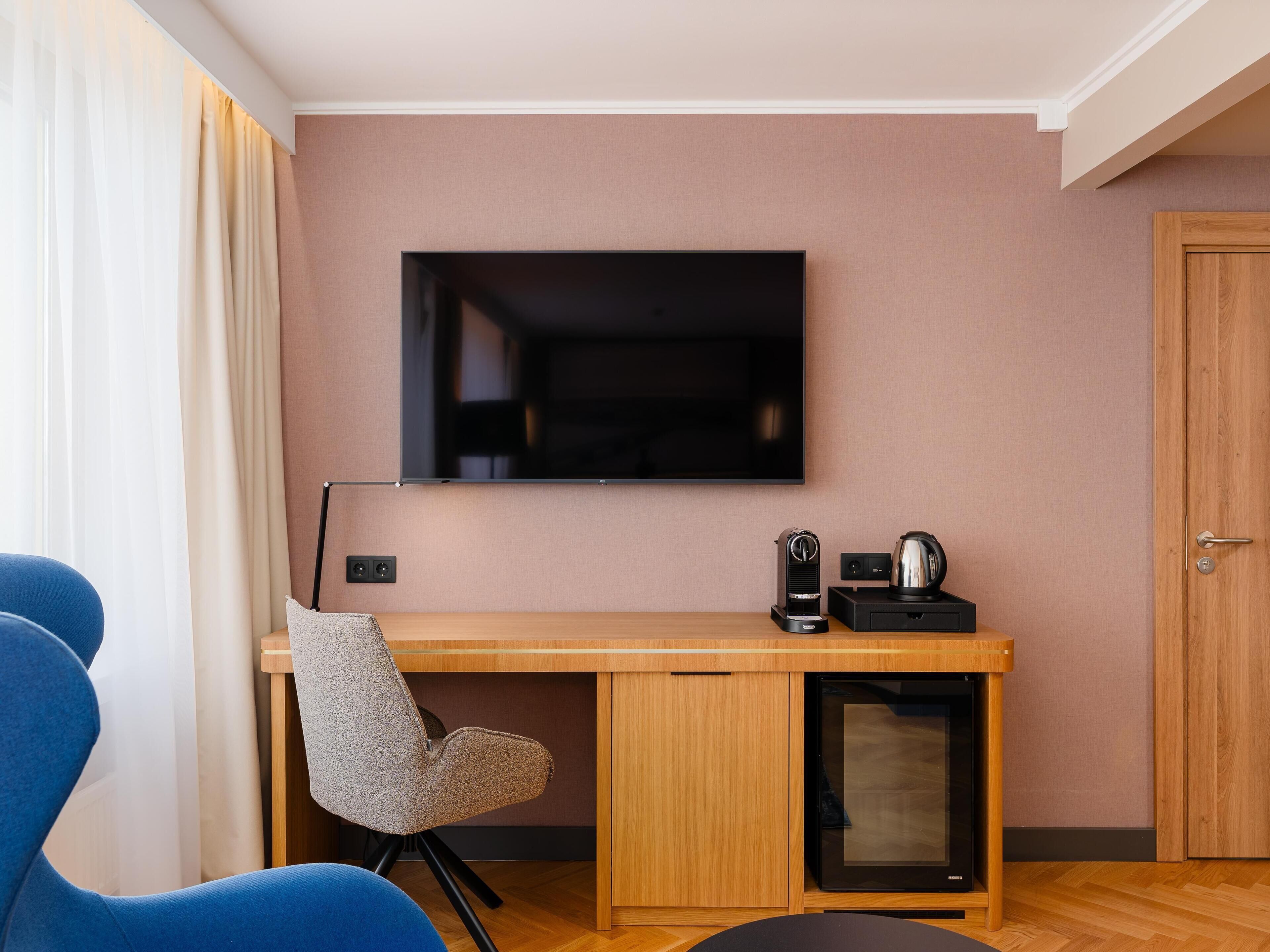 Moderni hotellihuone, jossa on puinen työpöytä, taulutelevisio, kahvinkeitin ja tuoli.