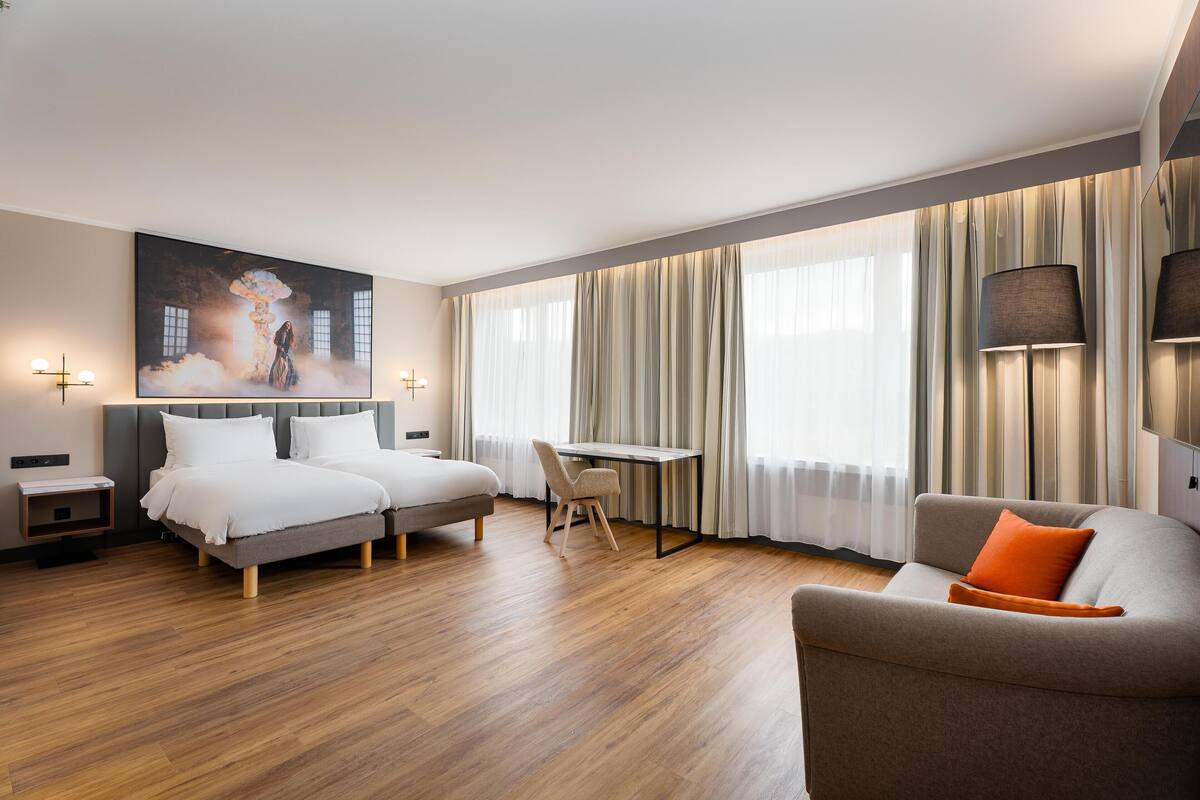 A modern hotel room with a bed, a sofa, a dining table, and a large painting on the wall.