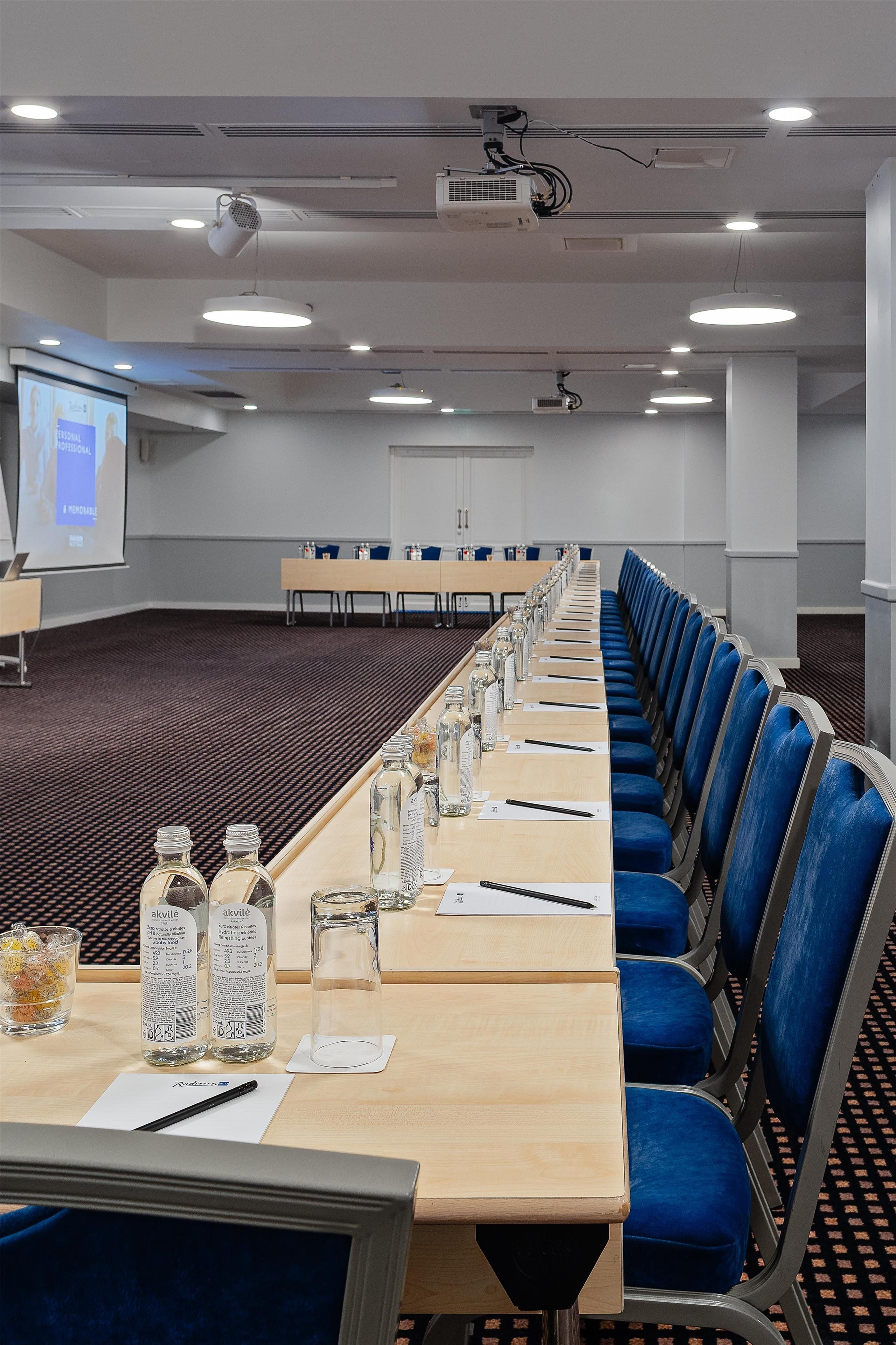 Un salón de conferencias con una larga mesa, sillas azules y una pantalla de proyector.