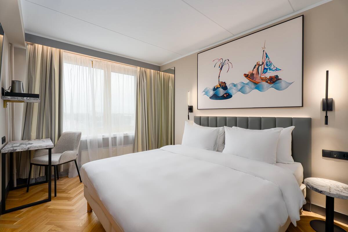A hotel room with a large bed, a desk, a chair, and a painting on the wall.