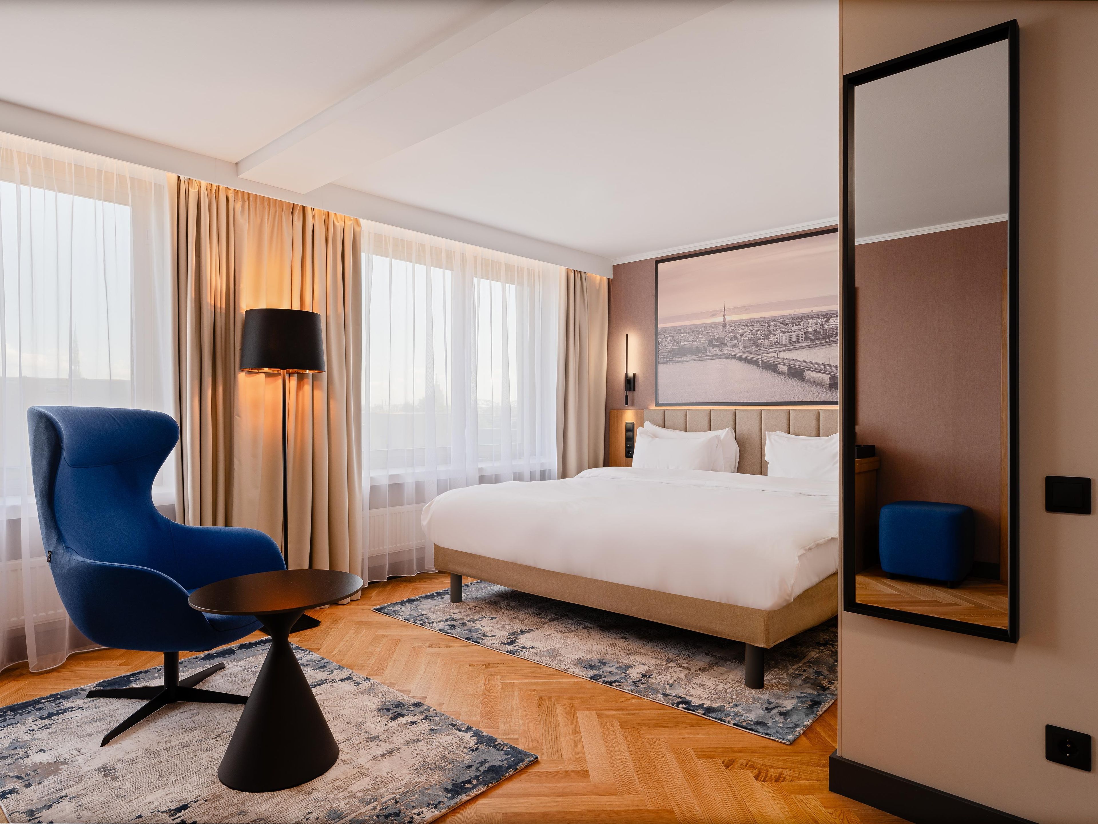 A hotel room with a bed, a chair, a lamp, and a large window with curtains.