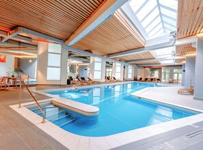 Una piscina interior con agua azul cristalina, terraza de madera y sillones de descanso.
