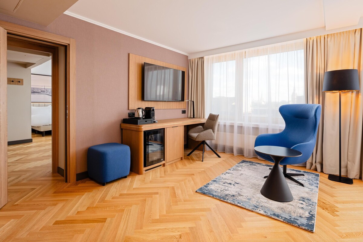 Een hotelkamer met een houten bureau, een televisie, een blauwe stoel, een lamp en uitzicht op de stad.