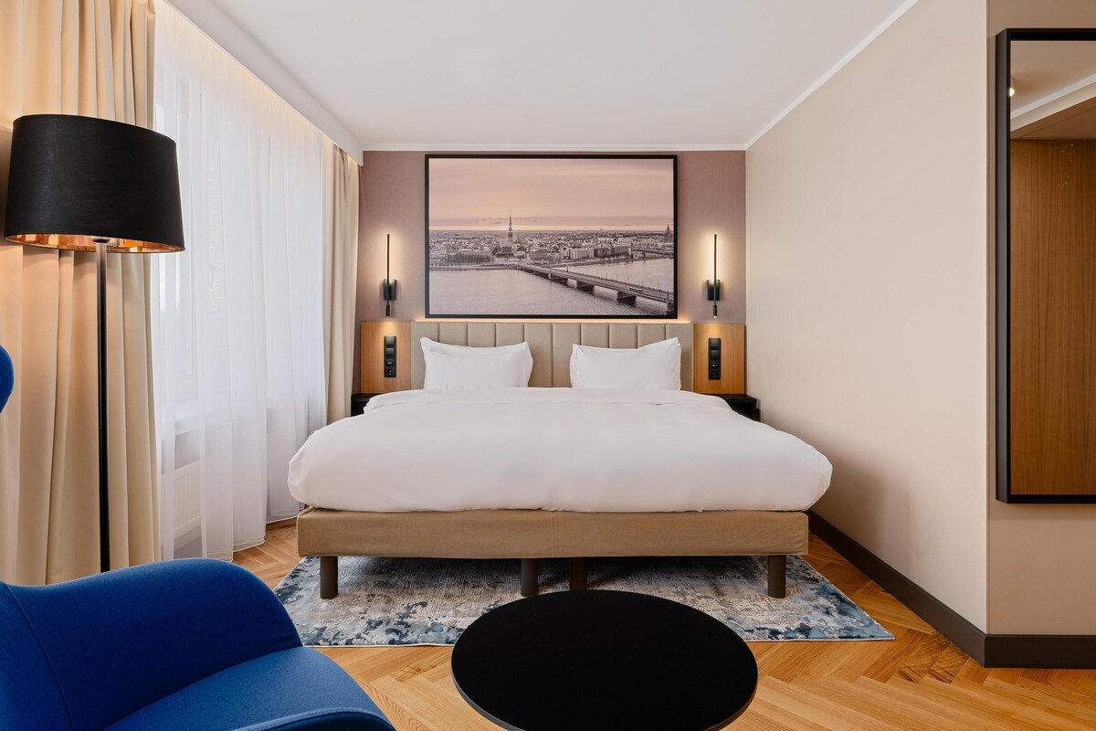 A hotel room with a bed, a nightstand, a lamp, a chair, and a large window with curtains.