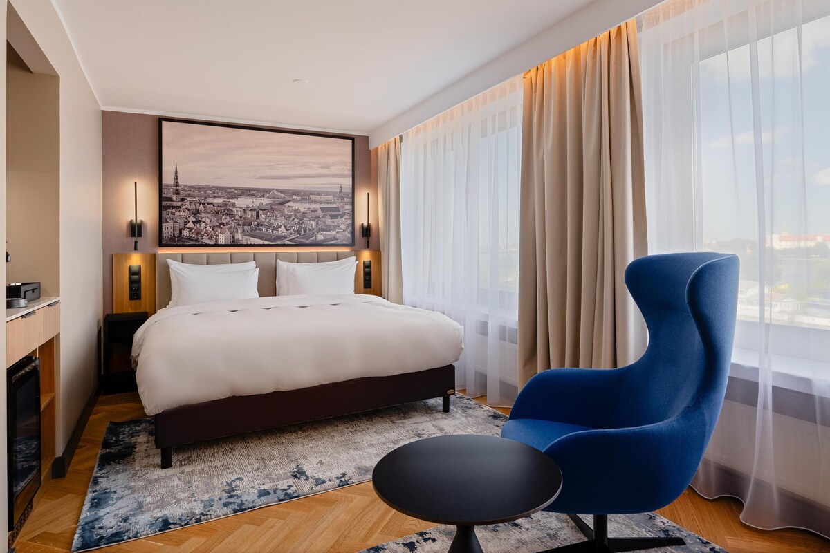 Een hotelkamer met een bed, een blauwe fauteuil, een klein tafeltje, een stadsgezicht schilderij en een groot raam met gordijnen.