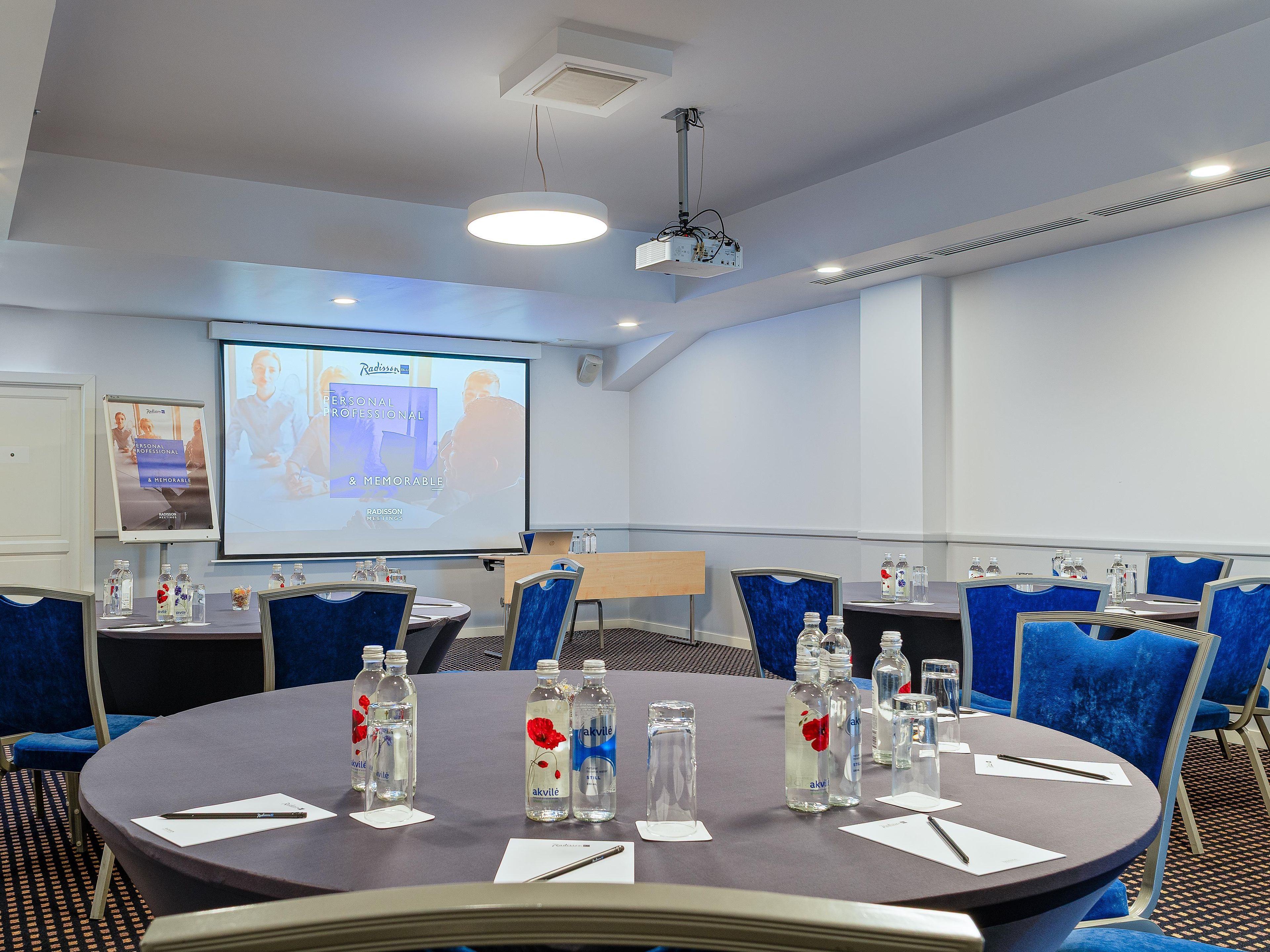 A conference room with a projector screen, tables with water bottles, and blue chairs.