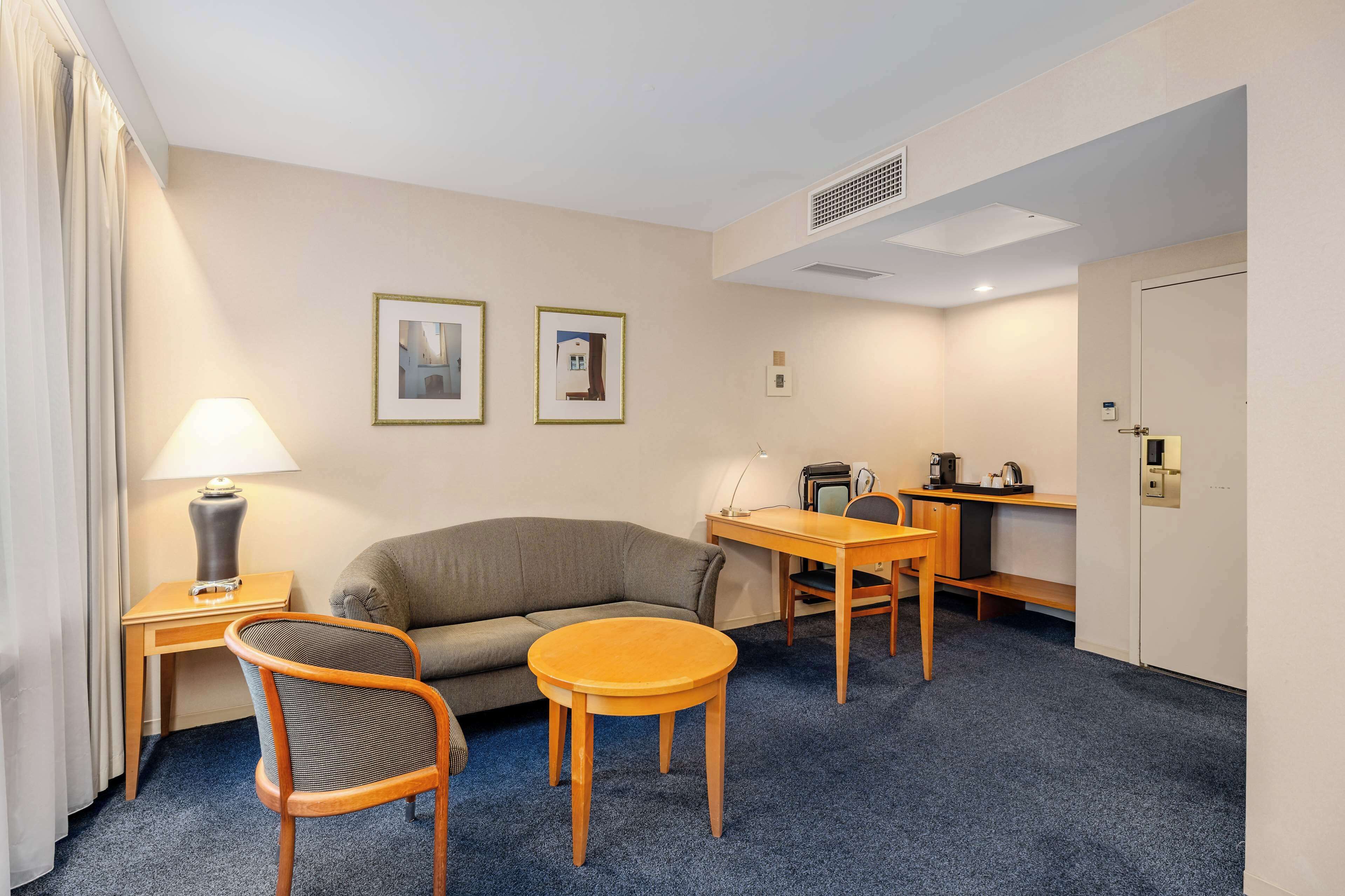 A hotel room with a sofa, a round table, a desk with a chair, a lamp, and two framed pictures on the wall.