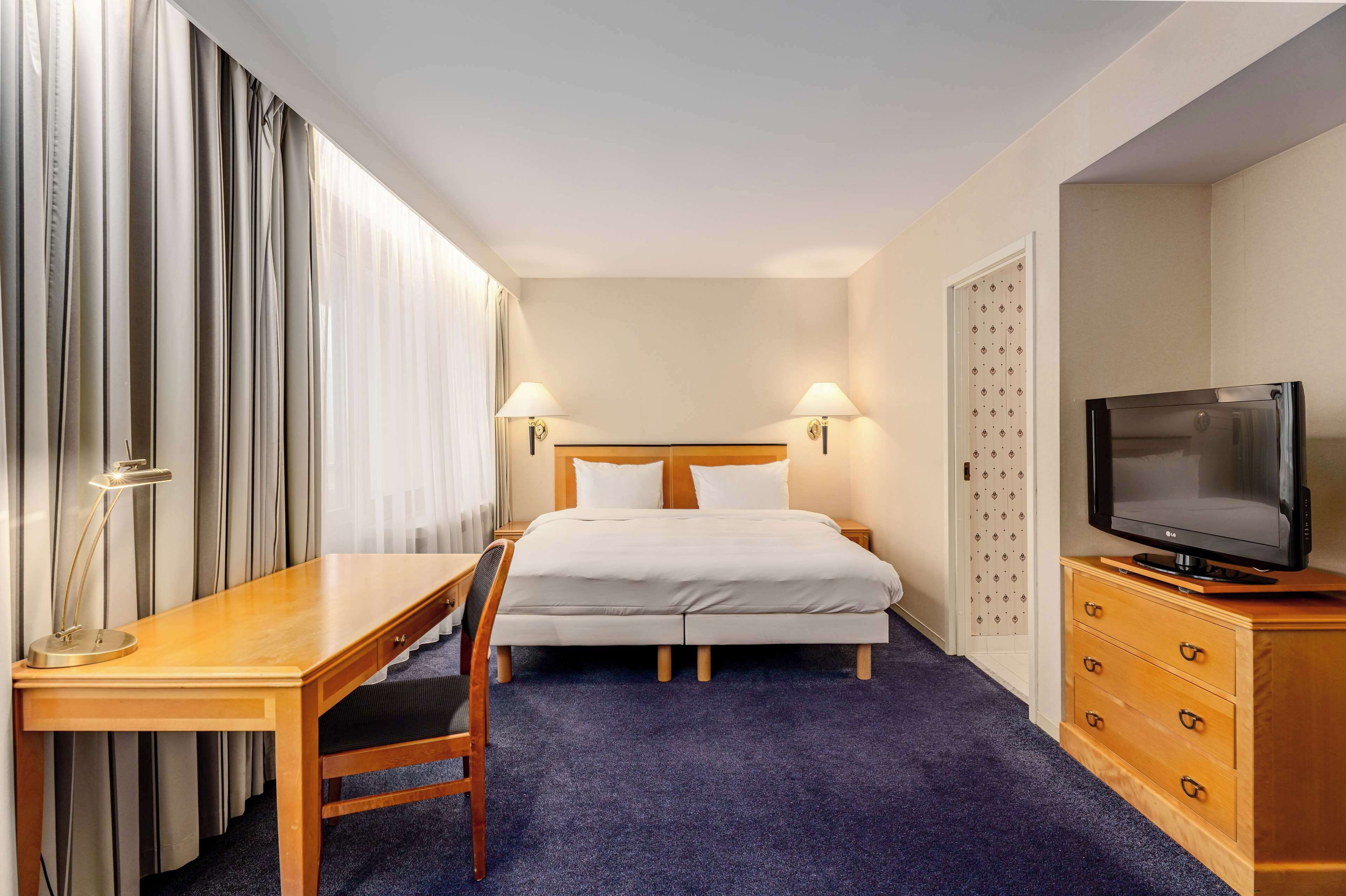 A hotel room with a bed, a desk, a chair, a TV, and a dresser.