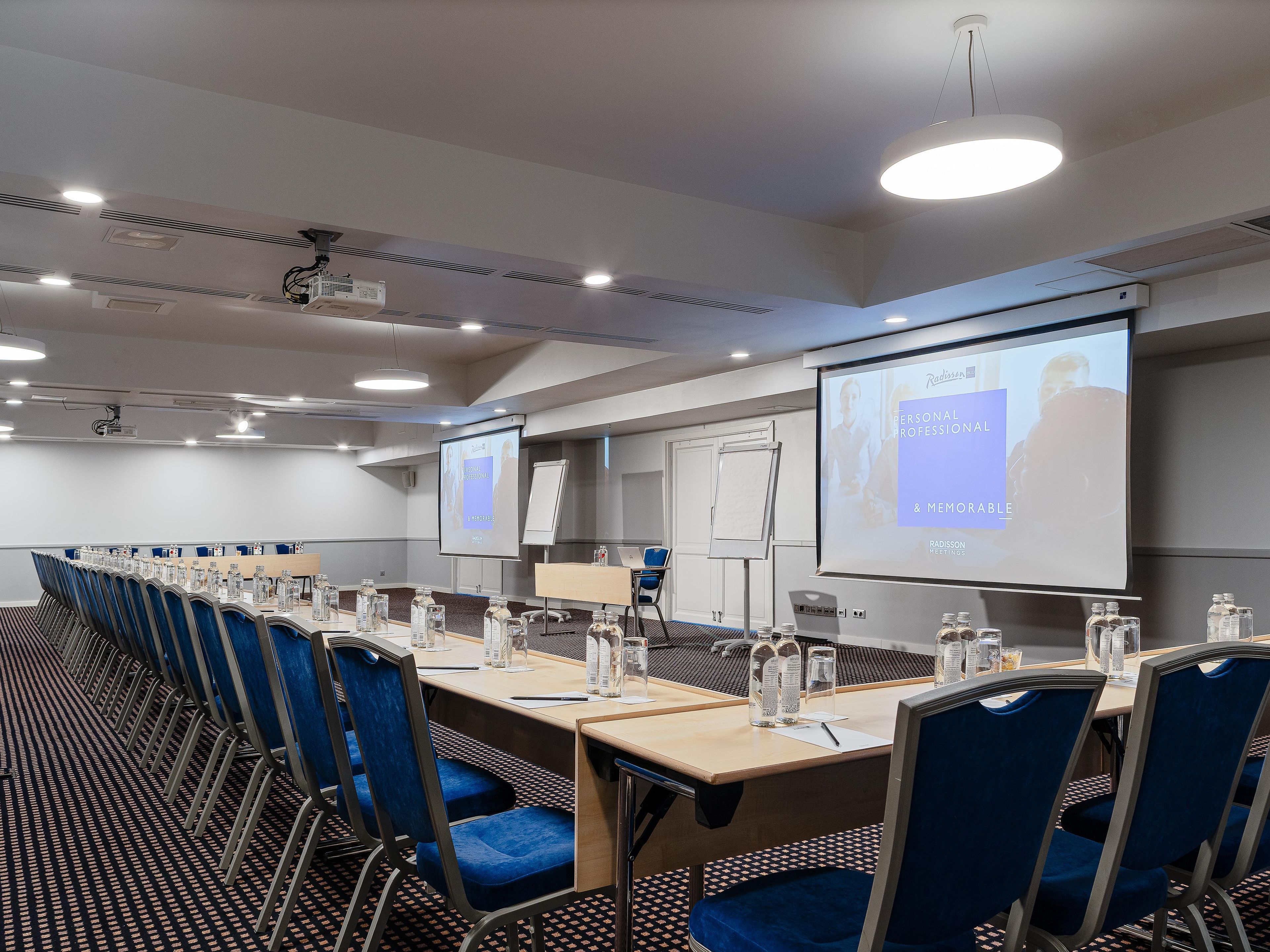 A conference room with a long table, blue chairs, a whiteboard, and a screen displaying a presentation.