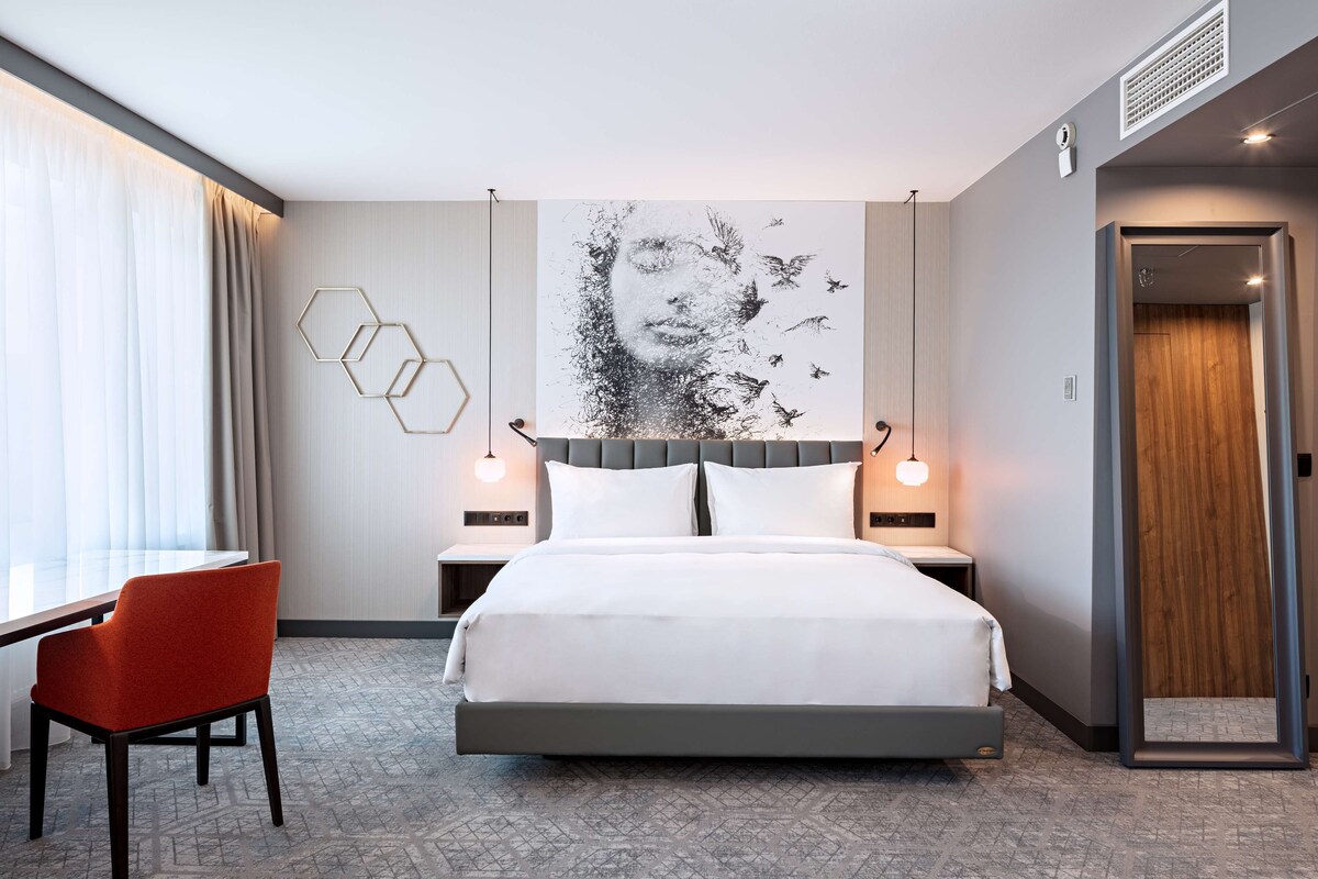 Een moderne hotelkamer met een groot bed, een rode stoel, een kunstwerk aan de muur en een geometrisch lamp.